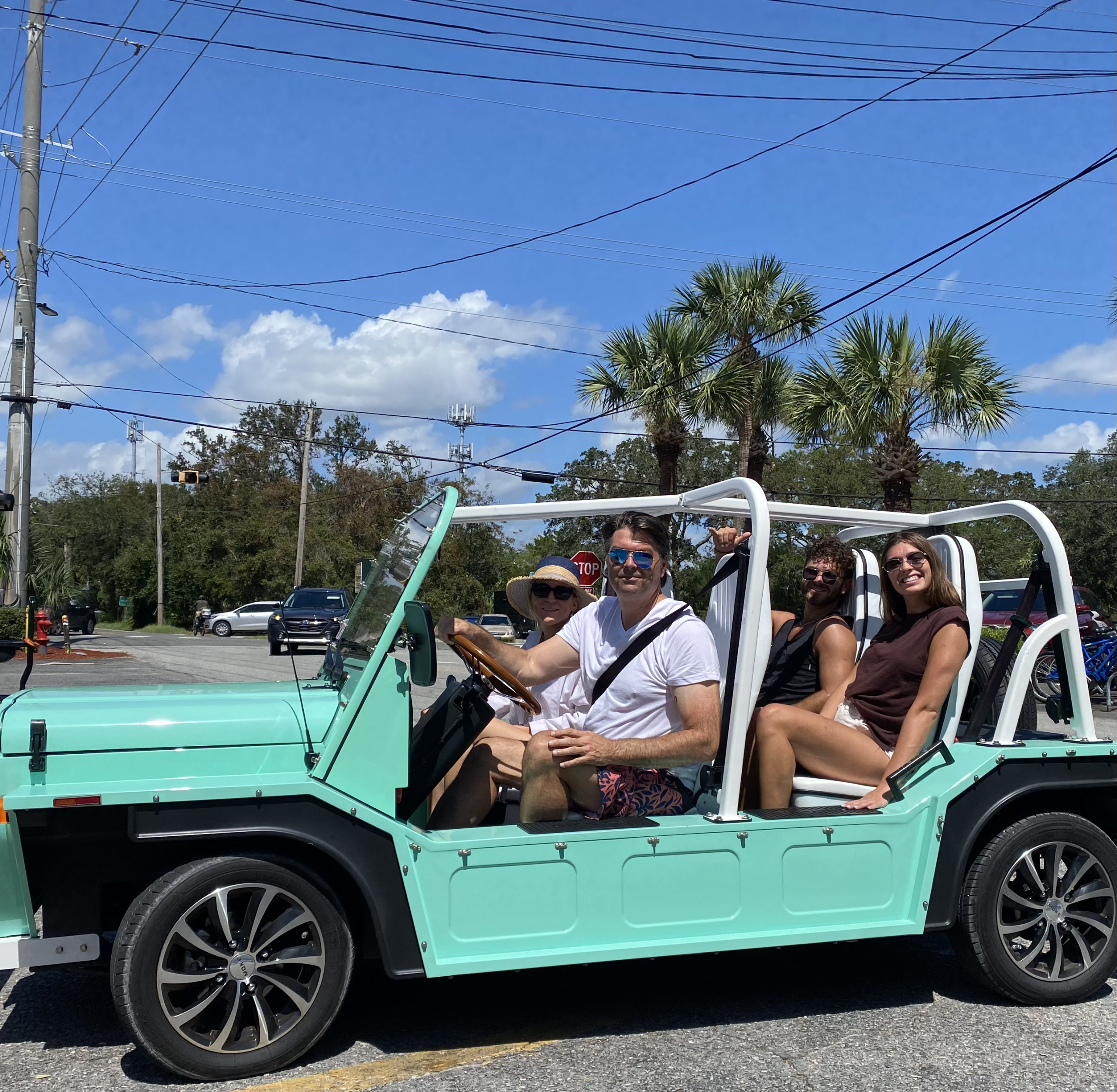 Four people sitting in a turquoise open-top vehicle on a sunny day, with palm trees and a stop sign in the background.