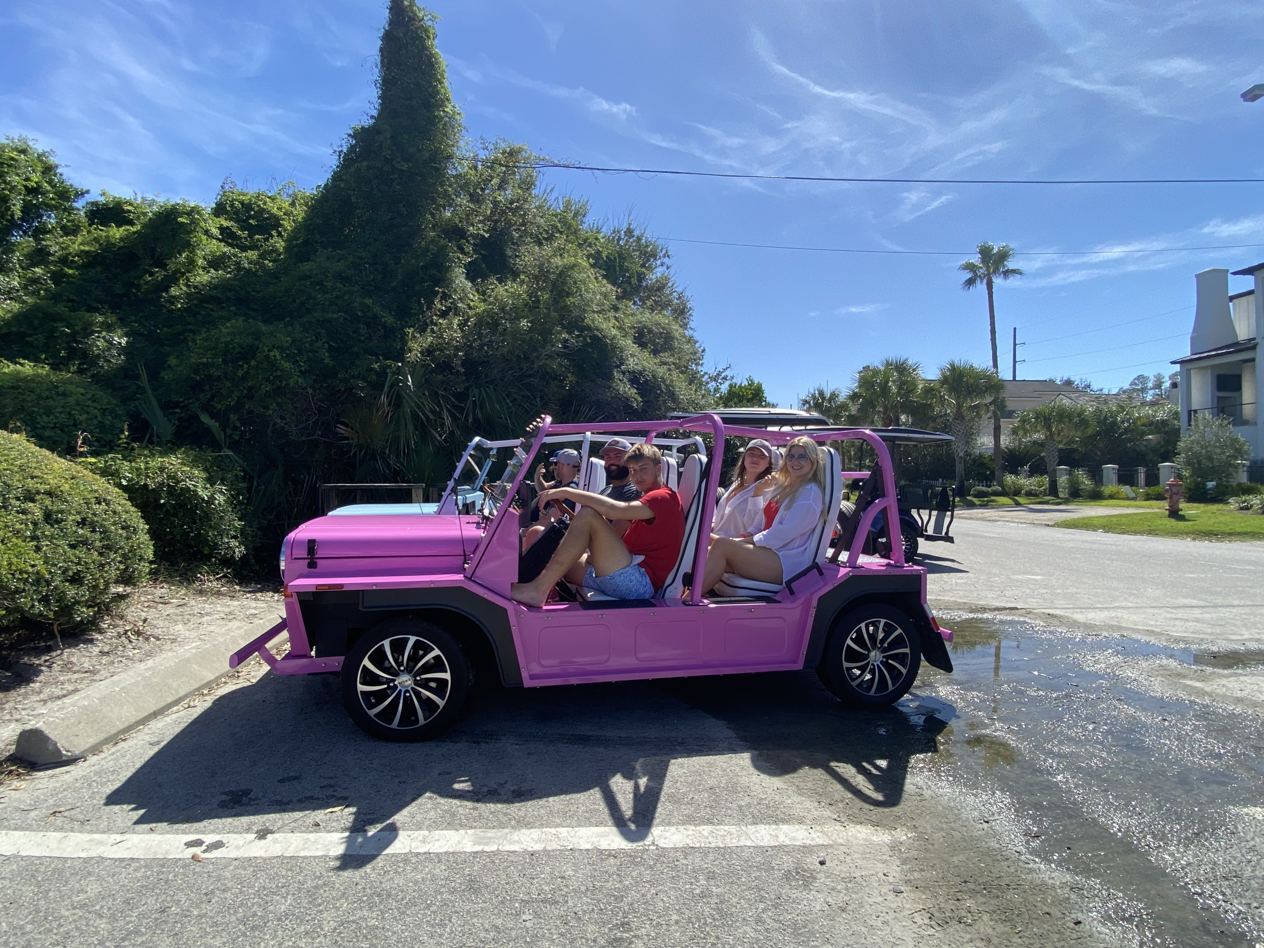 A pink off-road vehicle with four people inside, parked on a sunny street near greenery.